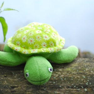 Op de afbeelding: Een handgemaakte groene pluche schildpad met een lachend gezicht en witte ogen. Het schild van de schildpad is gemaakt van lichtgroene stof met een wit en roze bloemenpatroon. De schildpad ligt op een verweerd houten oppervlak.