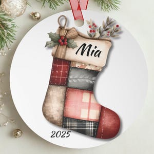 Op de afbeelding: Een ronde kerstversiering met een patchwork kousontwerp. De kous is versierd met verschillende patronen en kleuren, waaronder rood, grijs en beige. De naam "Mia" staat bovenaan en het jaar "2025" staat onderaan. Een rood lint is bevestigd om op te hangen.