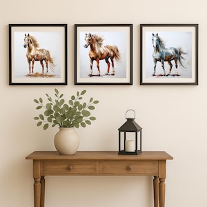 May include: Three framed horse art prints. The paintings depict horses in shades of brown, gold, and gray, with flowing manes and tails. The frames are black, and the artwork is displayed above a wooden console table.