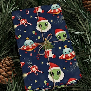 May include: A wrapped gift with navy blue wrapping paper featuring a repeating pattern of green aliens in Santa hats, UFOs, candy canes, and Christmas stockings. The gift is tied with twine and set against a backdrop of pine needles and a pine cone.