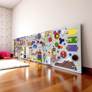 Può includere: Una grande tavola sensoriale grigia montata su un muro, con vari elementi interattivi colorati. Include ingranaggi, chiusure, interruttori e forme. La tavola è progettata per il gioco e l'apprendimento dei bambini, con un pavimento in legno in primo piano.