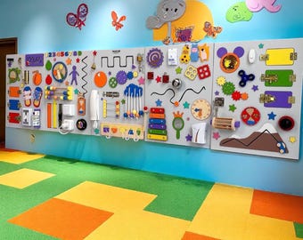 Könnte beinhalten: Eine sensorische Wand für Kinder mit verschiedenen interaktiven Elementen, darunter Zahnräder, Riegel und eine Uhr. Die Wand ist auf einer hellgrauen Oberfläche montiert und zeigt bunte Formen und Zahlen. Dekorative Schmetterlings- und Tierausstanzungen schmücken die blaue Wand darüber.