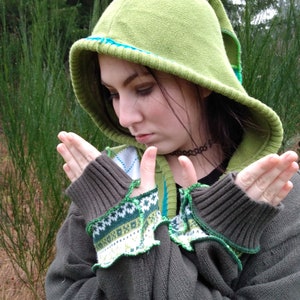 May include: A person wearing a green hooded sweater with a blue and white patterned trim. The sweater has a green and white patterned cuff. The person is holding their hands in front of them.
