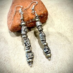 May include: Silver-toned dangle earrings with a unique, textured design. The earrings feature a series of beads and cylindrical elements, creating a layered effect. The earrings are displayed on a reddish-brown stone, showcasing their intricate details.