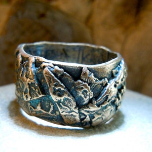 May include: A silver ring with a textured, abstract design. The ring has a wide band and a unique, organic shape.
