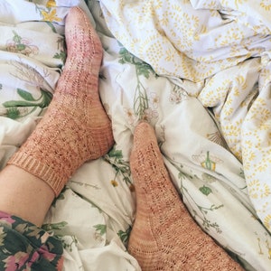 Könnte beinhalten: Zwei Paar beigefarbene Spitzensocken mit einem zarten Muster. Die Socken liegen auf einem weißen Bettüberwurf mit Blumenmuster.