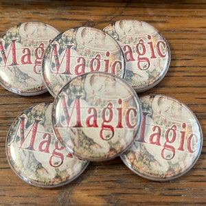 May include: A collection of five round buttons with the word "Magic" in red, stylized text. The background of each button features a faded, vintage-style illustration with muted colors. The buttons are arranged on a wooden surface.