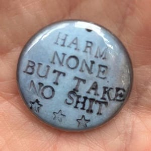 May include: A round, light blue button with the words "HARM NONE BUT TAKE NO SHIT" in dark letters. Four small star shapes are at the bottom of the button.