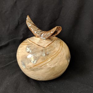 Può includere: Una scatola decorativa rotonda in ceramica con coperchio e manico curvo a forma di foglia. La scatola presenta un motivo marmorizzato beige e marrone con accenti argentati. Il manico è di colore marrone metallizzato.