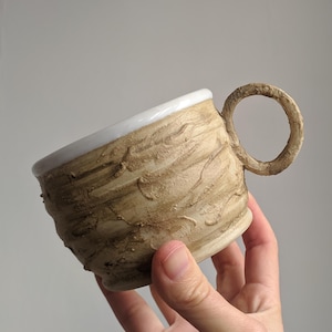 Può includere: Una tazza in ceramica fatta a mano con un esterno beige strutturato e un bordo interno bianco. La tazza presenta un manico arrotondato e un design unico e organico, perfetto per gustare caffè o tè.