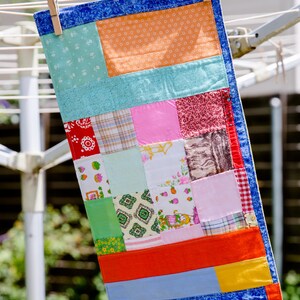 K&ouml;nnte beinhalten: Ein rechteckiger Patchwork-Quilt h&auml;ngt an einer W&auml;scheleine. Der Quilt zeigt Stoffquadrate in Blau, Orange, Rosa, Rot und Gr&uuml;n. Eine Holzklammer befestigt den Quilt an der Leine. Der Quilt ist etwa 76 cm lang.
