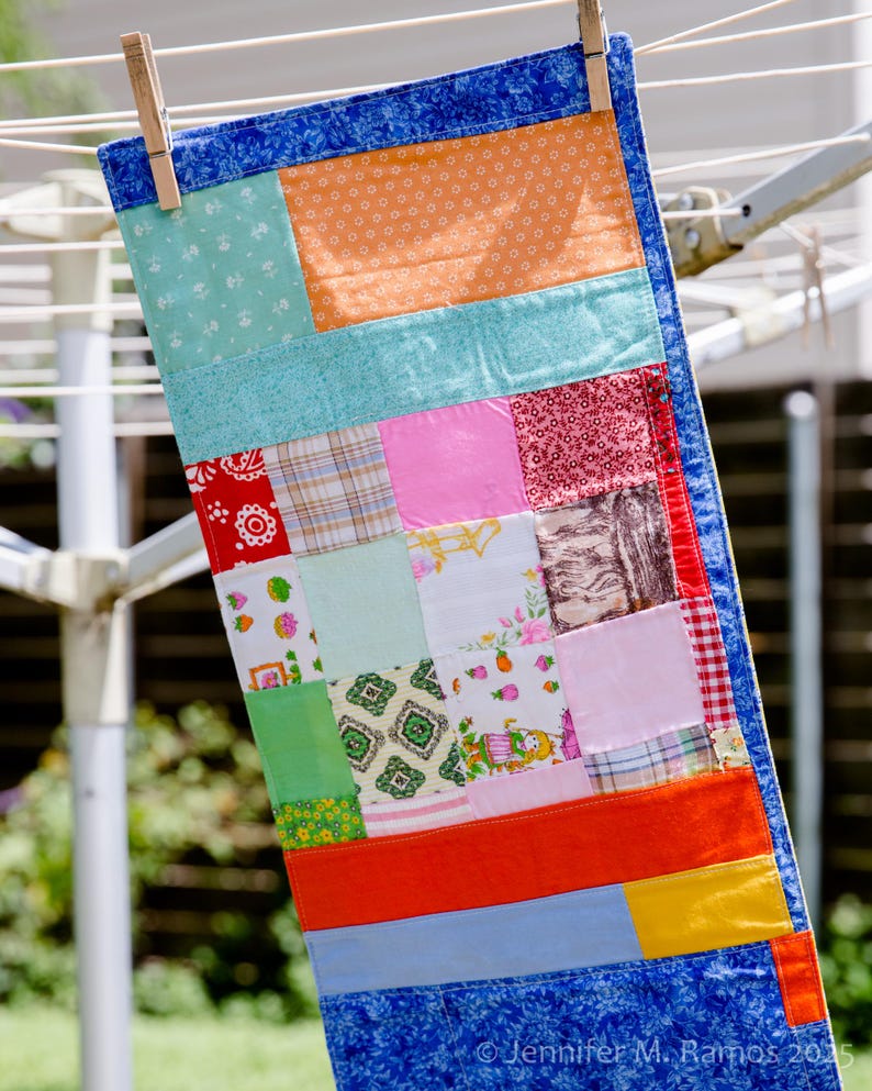 K&ouml;nnte beinhalten: Ein rechteckiger Patchwork-Quilt h&auml;ngt an einer W&auml;scheleine. Der Quilt zeigt Stoffquadrate in Blau, Orange, Rosa, Rot und Gr&uuml;n. Eine Holzklammer befestigt den Quilt an der Leine. Der Quilt ist etwa 76 cm lang.