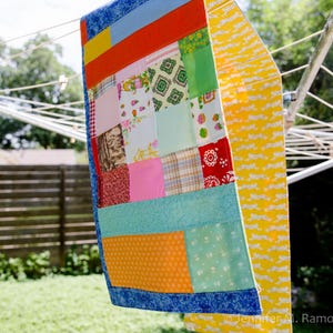 K&ouml;nnte beinhalten: Ein bunter Patchwork-Quilt mit blauem Rand h&auml;ngt an einer W&auml;scheleine. Der Quilt zeigt verschiedene Stoffmuster und -farben, darunter Rot, Orange, Rosa, Gr&uuml;n und Gelb. Ein gelber Stoff mit wei&szlig;em Muster h&auml;ngt ebenfalls an der W&auml;scheleine.