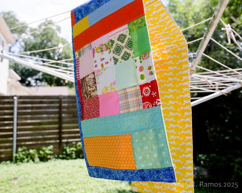 K&ouml;nnte beinhalten: Ein bunter Patchwork-Quilt mit blauem Rand h&auml;ngt an einer W&auml;scheleine. Der Quilt zeigt verschiedene Stoffmuster und -farben, darunter Rot, Orange, Rosa, Gr&uuml;n und Gelb. Ein gelber Stoff mit wei&szlig;em Muster h&auml;ngt ebenfalls an der W&auml;scheleine.