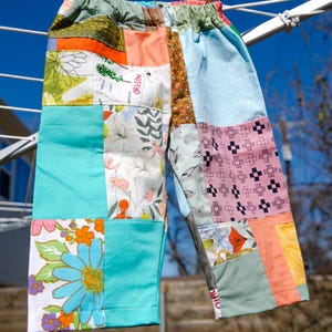 May include: Patchwork pants in various colors, including teal, orange, and floral patterns, hang on a clothesline. The pants are made of different fabric squares sewn together. The text "OREGON" is visible on one of the patches.