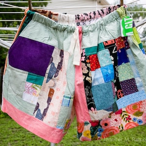 May include: Patchwork shorts with a drawstring waist, featuring a variety of fabric patterns and colors, including purple, pink, green, and floral prints. The shorts are hanging on a clothesline.