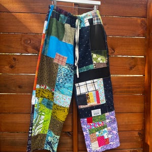 May include: Wide-leg pants constructed from a variety of multicolored fabric patches. The garment features an elastic waistband with a white drawstring, side pockets, and a rectangular white label on the right leg with black text.