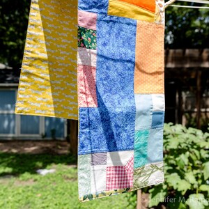 K&ouml;nnte beinhalten: Ein bunter Patchwork-Quilt h&auml;ngt an einer W&auml;scheleine. Der Quilt zeigt verschiedene Stoffmuster, darunter einen gelben Abschnitt mit wei&szlig;en Hunde-Silhouetten. Weitere Abschnitte zeigen blaue, orangefarbene, rosa und gemusterte Stoffe. Der Hintergrund ist unscharf.