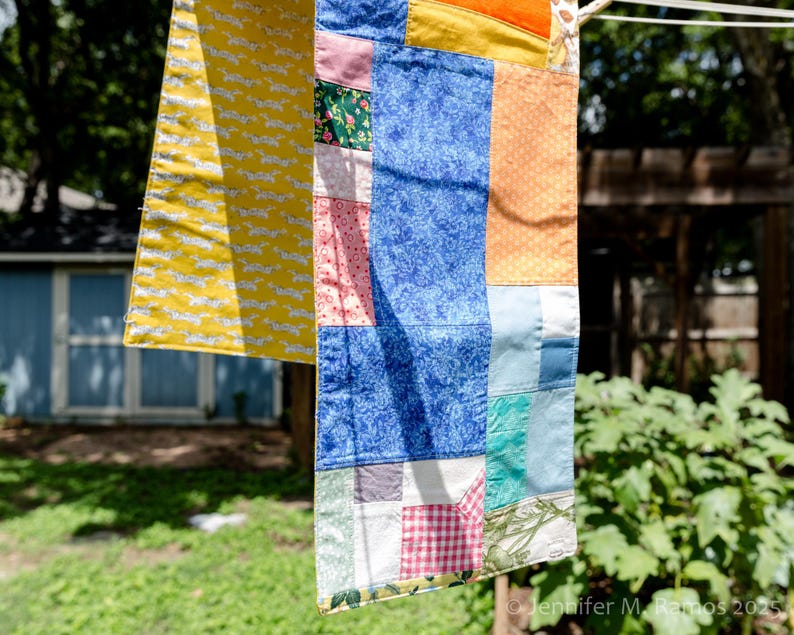 K&ouml;nnte beinhalten: Ein bunter Patchwork-Quilt h&auml;ngt an einer W&auml;scheleine. Der Quilt zeigt verschiedene Stoffmuster, darunter einen gelben Abschnitt mit wei&szlig;en Hunde-Silhouetten. Weitere Abschnitte zeigen blaue, orangefarbene, rosa und gemusterte Stoffe. Der Hintergrund ist unscharf.