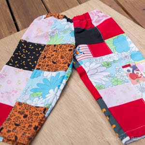 May include: A pair of pants featuring a patchwork design with various fabric patterns including orange and black Halloween motifs, floral prints in blue, pink, and white, and solid red and black panels. The pants have an elasticized waistband.