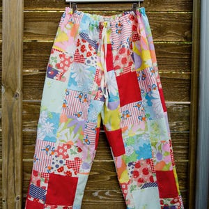 May include: Patchwork lounge pants featuring a colorful mix of fabric squares with various patterns including strawberries, flowers, and small figures. The pants have an elastic waistband with a white drawstring tie and a loose, wide-leg fit.