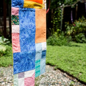 K&ouml;nnte beinhalten: Ein farbenfroher Patchwork-Quilt mit verschiedenen Stoffmustern und -farben, darunter blau floral, orange und rosa. Der Quilt h&auml;ngt im Freien, mit einem verschwommenen gr&uuml;nen Hintergrund.