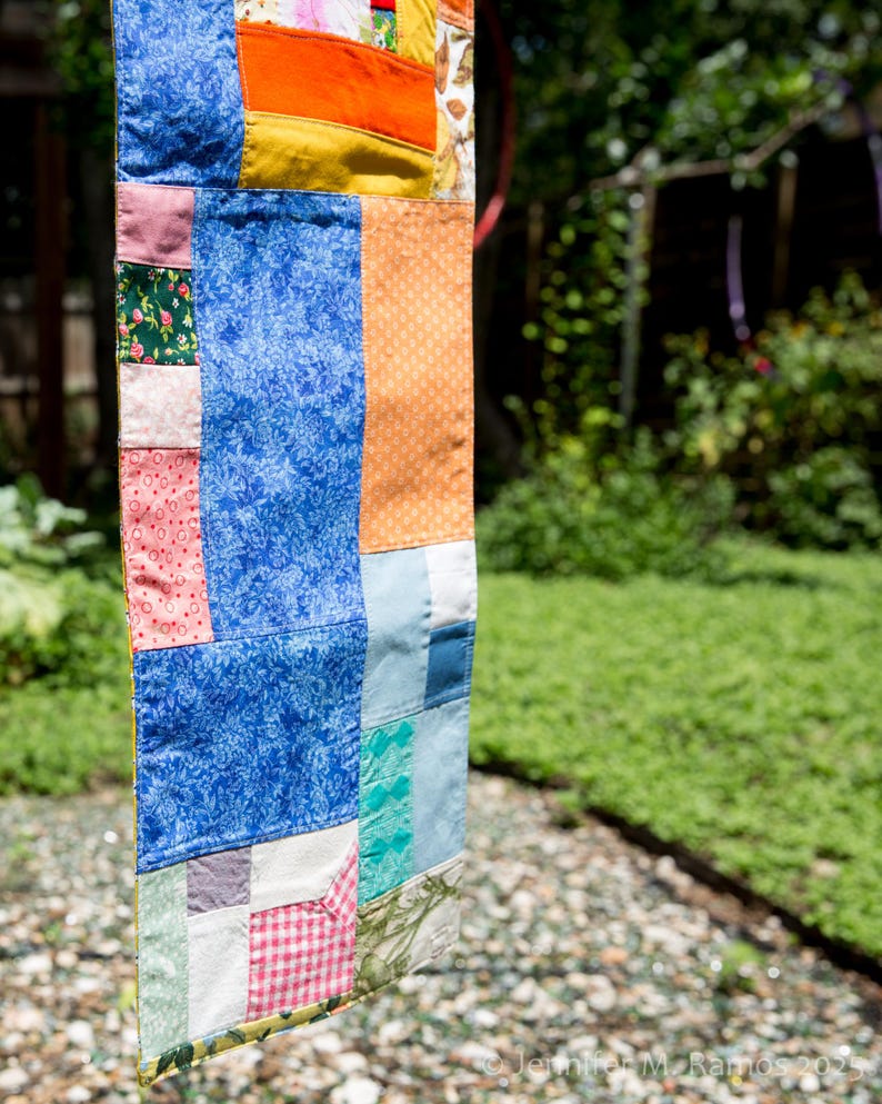 K&ouml;nnte beinhalten: Ein farbenfroher Patchwork-Quilt mit verschiedenen Stoffmustern und -farben, darunter blau floral, orange und rosa. Der Quilt h&auml;ngt im Freien, mit einem verschwommenen gr&uuml;nen Hintergrund.