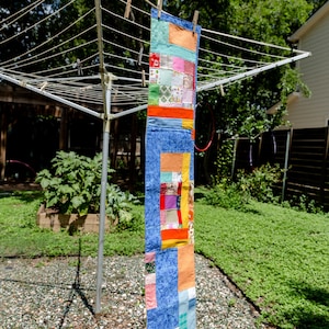 May include: A colorful, patchwork quilt banner hanging on a clothesline. The banner features a variety of fabric squares in shades of blue, orange, red, and green. The banner is secured with wooden clothespins, and the background shows a green yard.