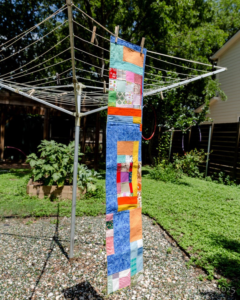 K&ouml;nnte beinhalten: Ein farbenfrohes Patchwork-Quilt-Banner, das an einer W&auml;scheleine h&auml;ngt. Das Banner besteht aus Stoffquadraten in Blau, Orange, Rot und Gr&uuml;n. Holzklammern befestigen das Banner. Im Hintergrund ist ein gr&uuml;ner Garten zu sehen.