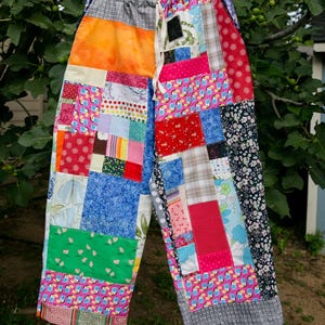 May include: Patchwork pants featuring a wide-leg silhouette and an elastic waistband with a white drawstring. The garment is constructed from various fabric squares in diverse colors, including bright pink, blue, red, green, and orange, with patterns such as toucans, florals, and plaid.