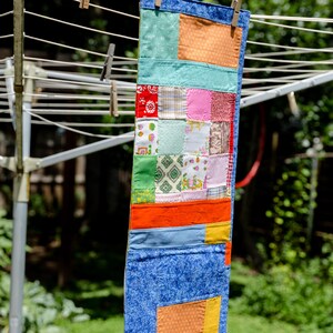 K&ouml;nnte beinhalten: Ein farbenfrohes Patchwork-Quilt h&auml;ngt an einer W&auml;scheleine. Der Quilt besteht aus verschiedenen Stoffen in Blau-, Orange-, Gr&uuml;n-, Rot- und Rosat&ouml;nen. Er wird mit Holzw&auml;scheklammern befestigt. Der Hintergrund ist eine verschwommene Ansicht von gr&uuml;nem Laub.