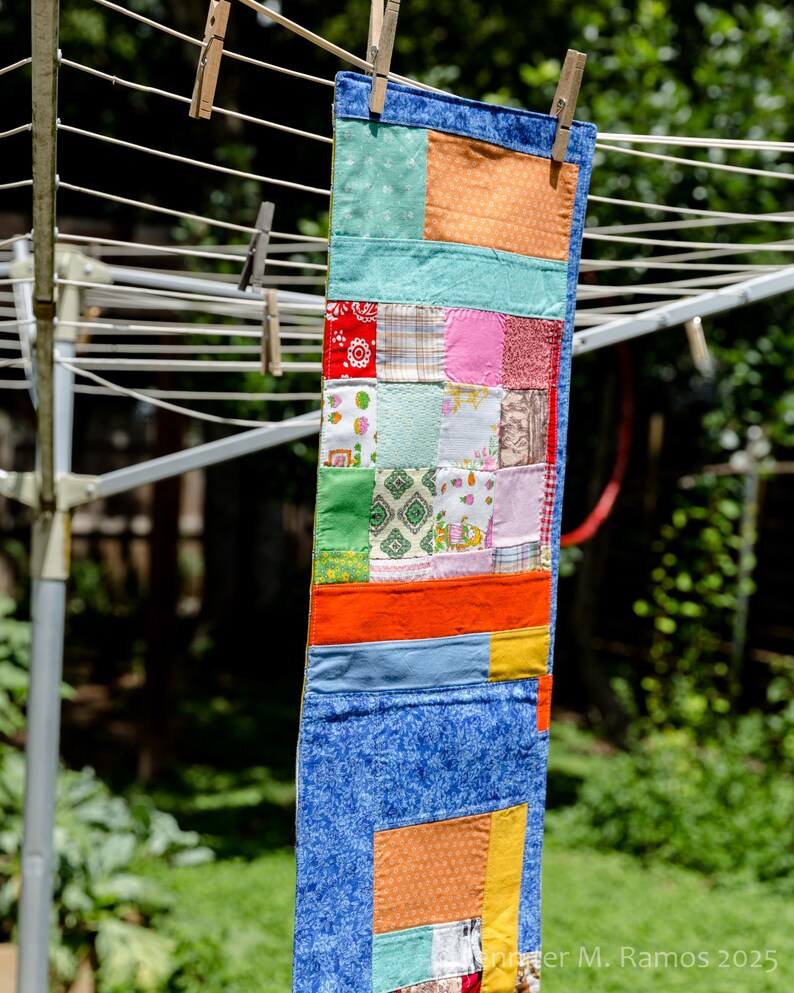 K&ouml;nnte beinhalten: Ein farbenfrohes Patchwork-Quilt h&auml;ngt an einer W&auml;scheleine. Der Quilt besteht aus verschiedenen Stoffen in Blau-, Orange-, Gr&uuml;n-, Rot- und Rosat&ouml;nen. Er wird mit Holzw&auml;scheklammern befestigt. Der Hintergrund ist eine verschwommene Ansicht von gr&uuml;nem Laub.