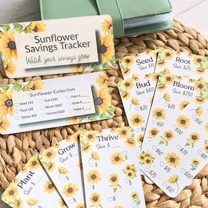May include: A set of sunflower-themed savings trackers and a green card holder. The trackers feature sunflower illustrations and text like "Sunflower Savings Tracker" and "Watch your savings grow." The cards are arranged on a woven mat.