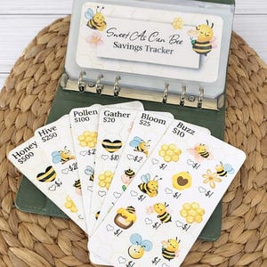 May include: A green binder with a "Sweet As Can Bee Savings Tracker" card. The binder contains cards with bee illustrations and text. The cards are for tracking savings goals. The background is a woven, light brown surface.