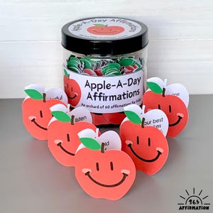以下が含まれることがあります： 「Apple-A-Day Affirmations」と書かれたラベルが付いた黒い蓋の、リンゴ型の肯定カードが入った透明な瓶。 瓶の前には、笑顔の顔と肯定的な言葉が書かれた赤いリンゴ型のカードがいくつか配置されています。