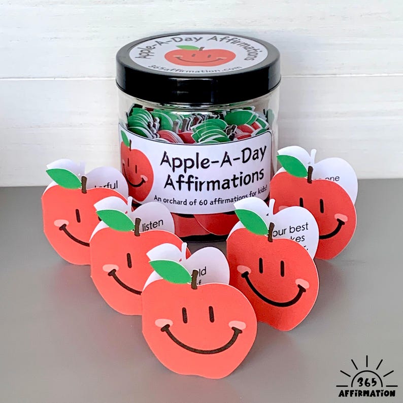 以下が含まれることがあります： 「Apple-A-Day Affirmations」と書かれたラベルが付いた黒い蓋の、リンゴ型の肯定カードが入った透明な瓶。 瓶の前には、笑顔の顔と肯定的な言葉が書かれた赤いリンゴ型のカードがいくつか配置されています。