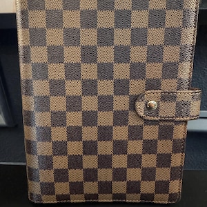 May include: Brown and tan checkered pattern notebook cover with a snap closure. The cover has a classic, geometric design and a rectangular shape. The snap closure is a small, round gold-colored button. The notebook cover is made of a textured material.
