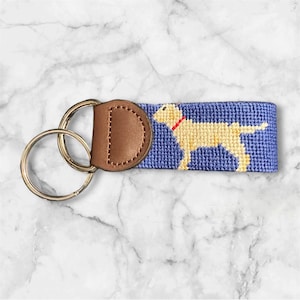 Puede incluir: Un llavero azul y blanco con un diseño de Labrador Retriever amarillo. El llavero tiene un lazo de cuero marrón y dos llaveros de plata.