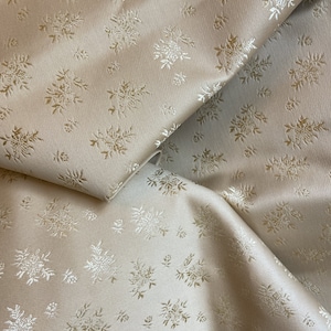 May include: A close-up of a beige fabric with a white floral pattern. The fabric is folded in half, showing the pattern on both sides.