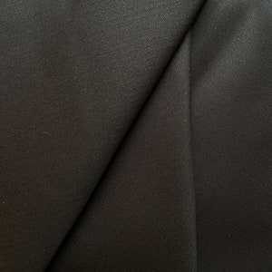 Peut inclure: Gros plan sur un tissu noir avec un motif de tissage en chevrons subtil. Le tissu est plié, créant des lignes diagonales et des ombres qui mettent en valeur la texture et la profondeur du matériau. La couleur sombre suggère un textile formel ou polyvalent.
