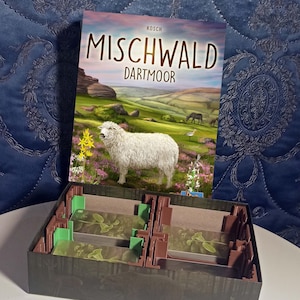 Può includere: Scatola di gioco da tavolo "MISCHWALD DARTMOOR" con un'illustrazione di pecora. La scatola è aperta, rivelando un interno marrone e verde con divisori. La scatola del gioco è su una superficie bianca.