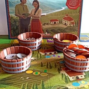 Peut inclure: Un jeu de société appelé "Vticulture: Essential Edition" avec une boîte représentant un couple debout dans un vignoble. Le plateau de jeu montre un vignoble avec une charrette, un bâtiment et un champ avec une rangée de jetons jaunes, verts et bruns. Il y a quatre barils en bois avec des jetons à l'intérieur.