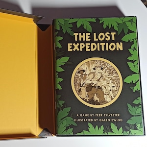 Puede incluir: Caja de juego de mesa abierta titulada "THE LOST EXPEDITION". La caja es de color verde oscuro con un diseño de hojas de la jungla y una ilustración circular de exploradores. El interior de la caja es amarillo.