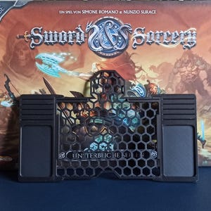 Könnte beinhalten: Ein schwarzer Plastik-Spieleorganizer mit einem Wabenmuster und zwei Steckplätzen für Karten. Der Organizer ist mit "Unterbliche Seiten" beschriftet.