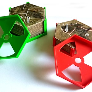 Könnte beinhalten: Zwei Sätze von Spielkartenhaltern, einer grün und einer rot, jeweils mit sechseckiger Form und einem Ausschnitt mit Strahlensymbol. Die Halter befinden sich neben Stapeln von Spielkarten mit einem Kartendesign.