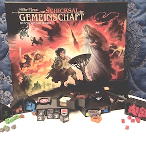 Könnte beinhalten: Ein Brettspielkasten mit dem Titel "Herr der Ringe - Das Schicksal der Gemeinschaft" und verschiedene Spielteile. Das Box-Art zeigt eine Fantasy-Szene mit Figuren und einem feurigen Hintergrund. Würfel, Spielsteine und Aufbewahrungsbehälter sind sichtbar.