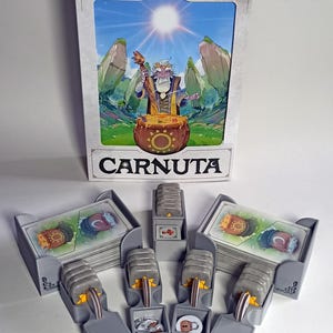 Puede incluir: Juego de mesa "CARNUTA" con una caja que presenta una ilustración de un mago y un caldero. El juego incluye cartas y componentes de plástico gris. El nombre de los creadores del juego también se muestra.