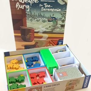 Könnte beinhalten: Ein Brettspiel-Box-Set mit dem Titel "Die Weiße Burg" und "Die Tee-Zeremonie" wird gezeigt. Die Box enthält verschiedene Spielsteine in Gelb, Blau, Grün und Rot, sowie Karten und einen grünen Trenner. Die Box hat auch den Text "3 RING CIRCUS".