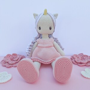 K&ouml;nnte beinhalten: Geh&auml;kelte Einhorn-Puppe mit hellrosa Kleid, wei&szlig;em Haar und gelbem Horn. Die Puppe hat gro&szlig;e, dunkle Augen und sitzt auf einer rosa Oberfl&auml;che. Geh&auml;kelte Blumen und Wolkenformen sind ebenfalls vorhanden.