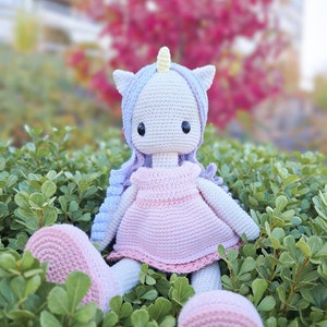 K&ouml;nnte beinhalten: Handgefertigte H&auml;kel-Einhorn-Puppe mit hellblauer und lavendelfarbener M&auml;hne, gelbem Horn und rosa Kleid. Die Puppe sitzt inmitten von gr&uuml;nem Laub, mit einem passenden rosa Fu&szlig; und einem runden Ball.
